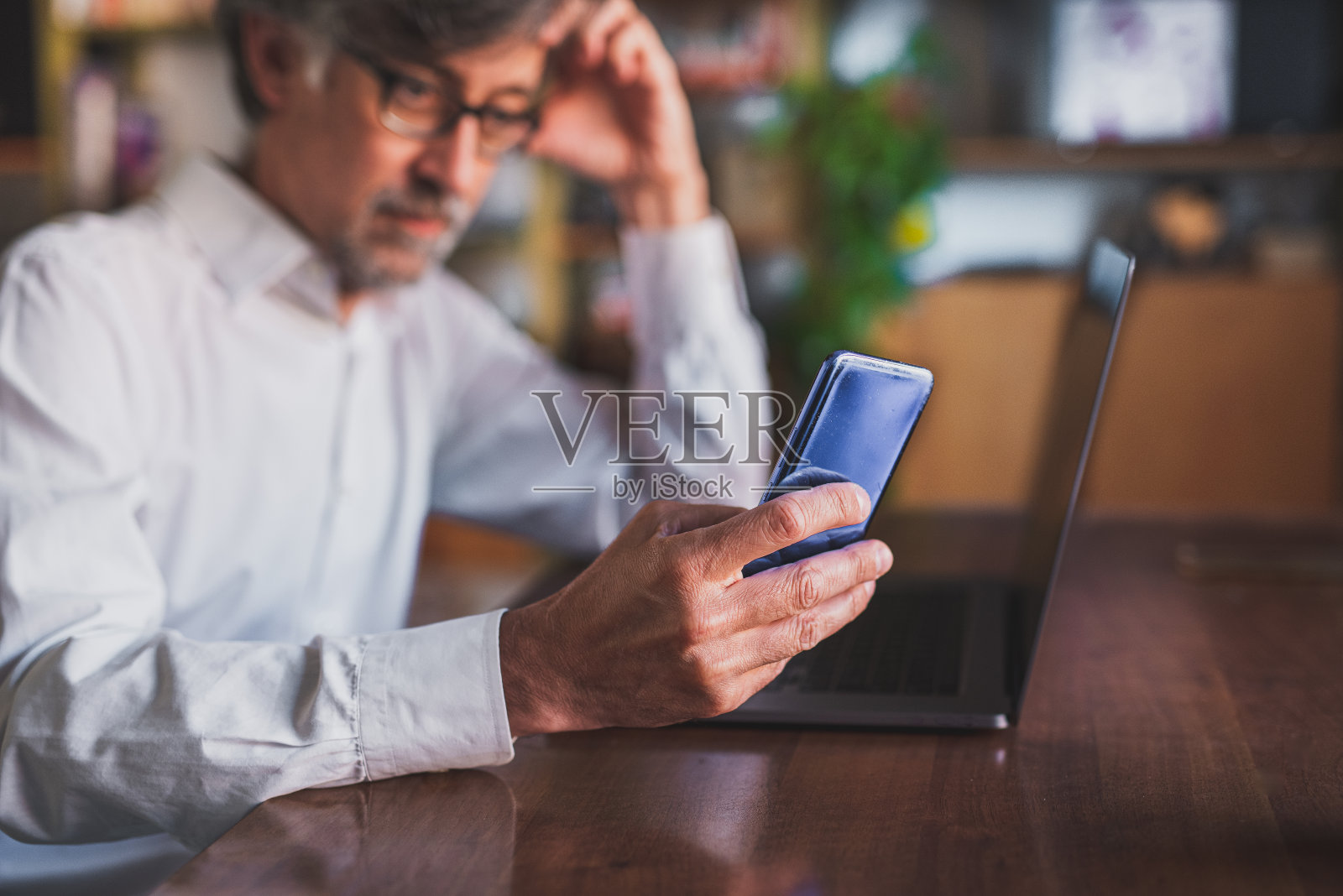 深思熟虑的商人戴着眼镜坐在笔记本电脑前，以一种智能的方式在家工作，使用智能手机连接和发送信息，专注于手和智能手机，模糊的背景照片摄影图片