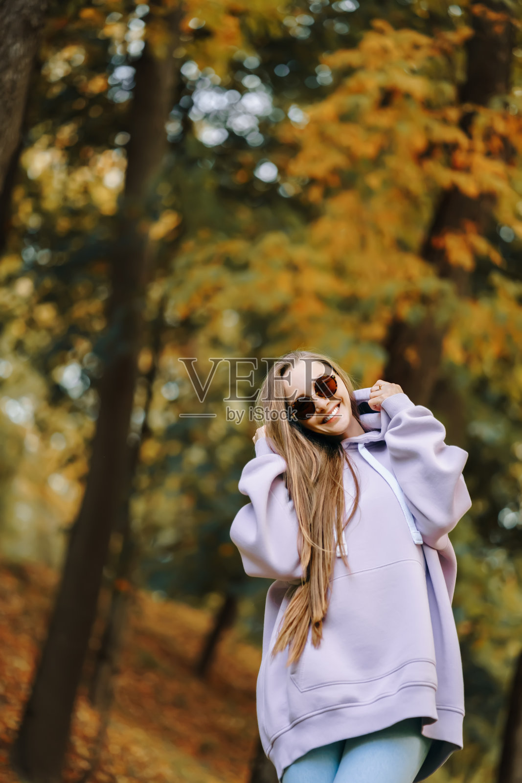 在阳光明媚的秋日，穿着粉色帽衫和牛仔裤的年轻女子在公园里。季节与人的概念照片摄影图片