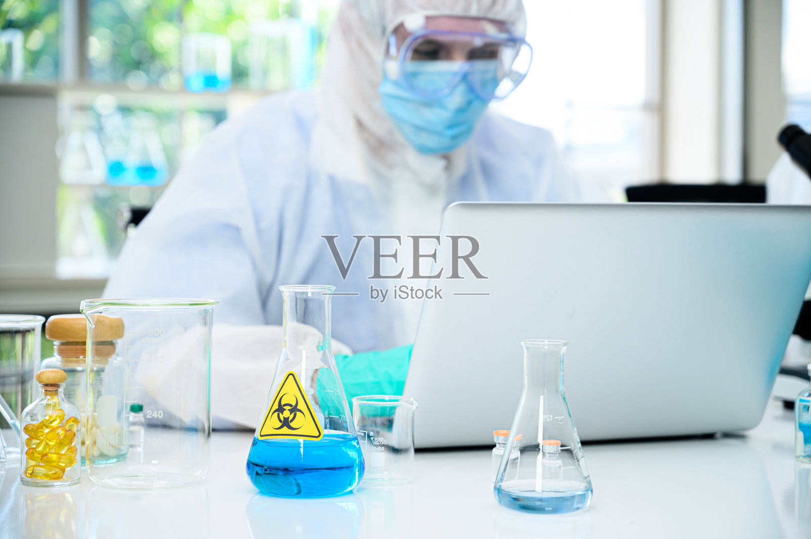 桌子上放着装有蓝色液体的实验室设备，背景是穿着防护服的男性科学家在实验室里用笔记本电脑进行研究。照片摄影图片