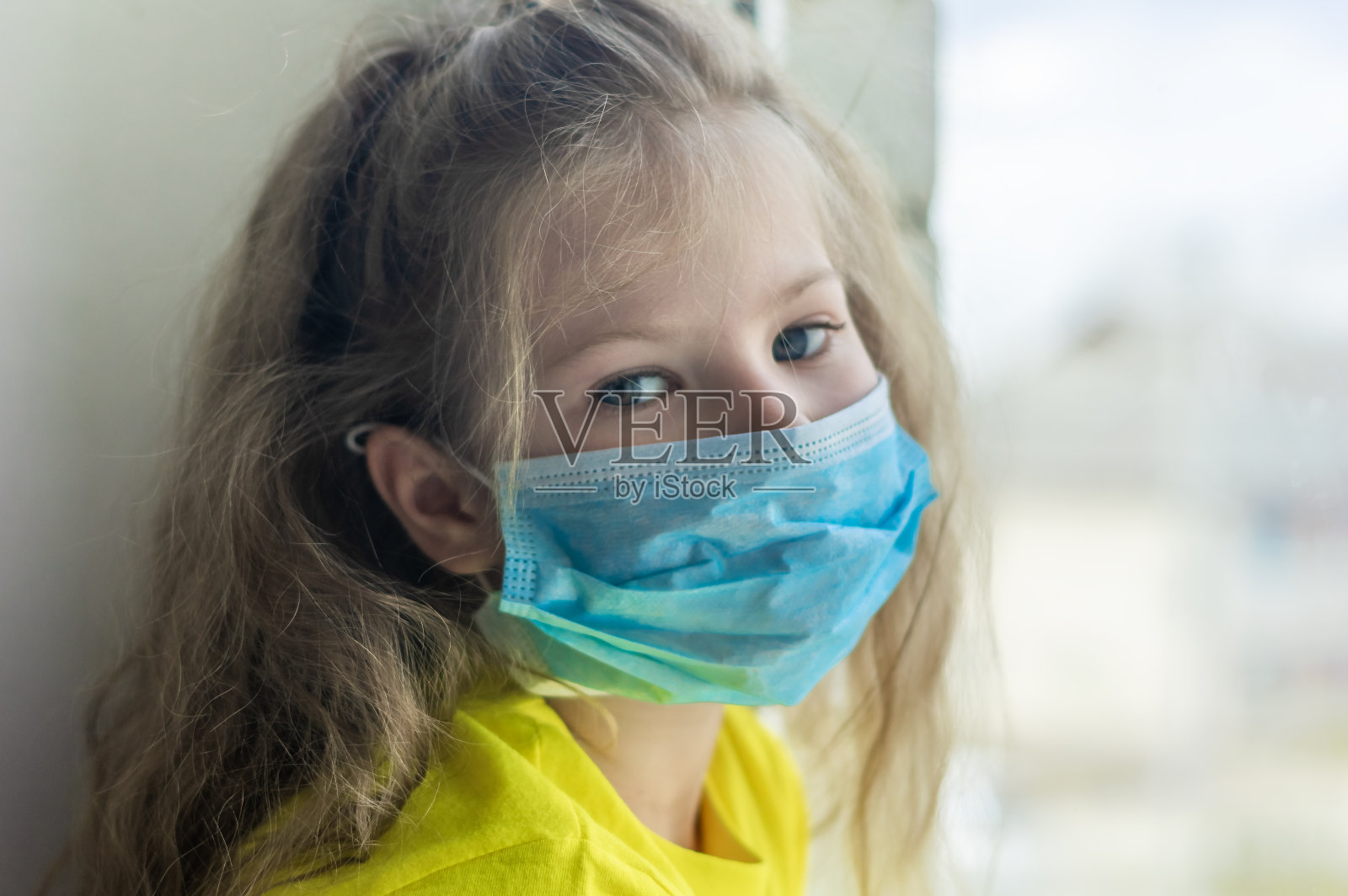 一位戴口罩的学龄前女孩在被冠状病毒隔离期间在家自我隔离的肖像。看着镜头难过。闭合抑郁的概念。照片摄影图片