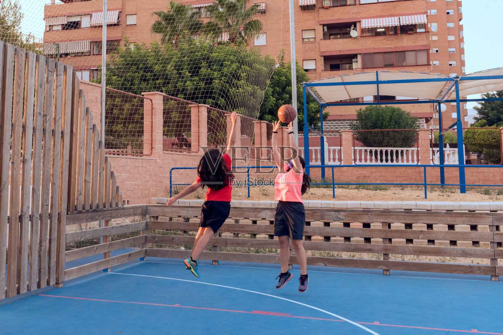 两个年轻的女孩在城市的街道球场打篮球，一个投掷，另一个试图阻挡。照片摄影图片