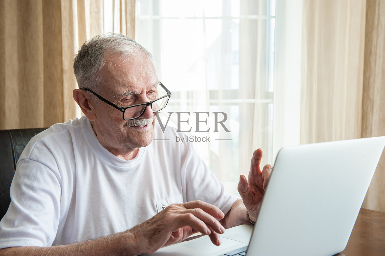 一个老人坐在家里的笔记本电脑前。一位白发苍苍、戴眼镜的老人在家使用笔记本电脑工作。老年人沟通的概念照片摄影图片