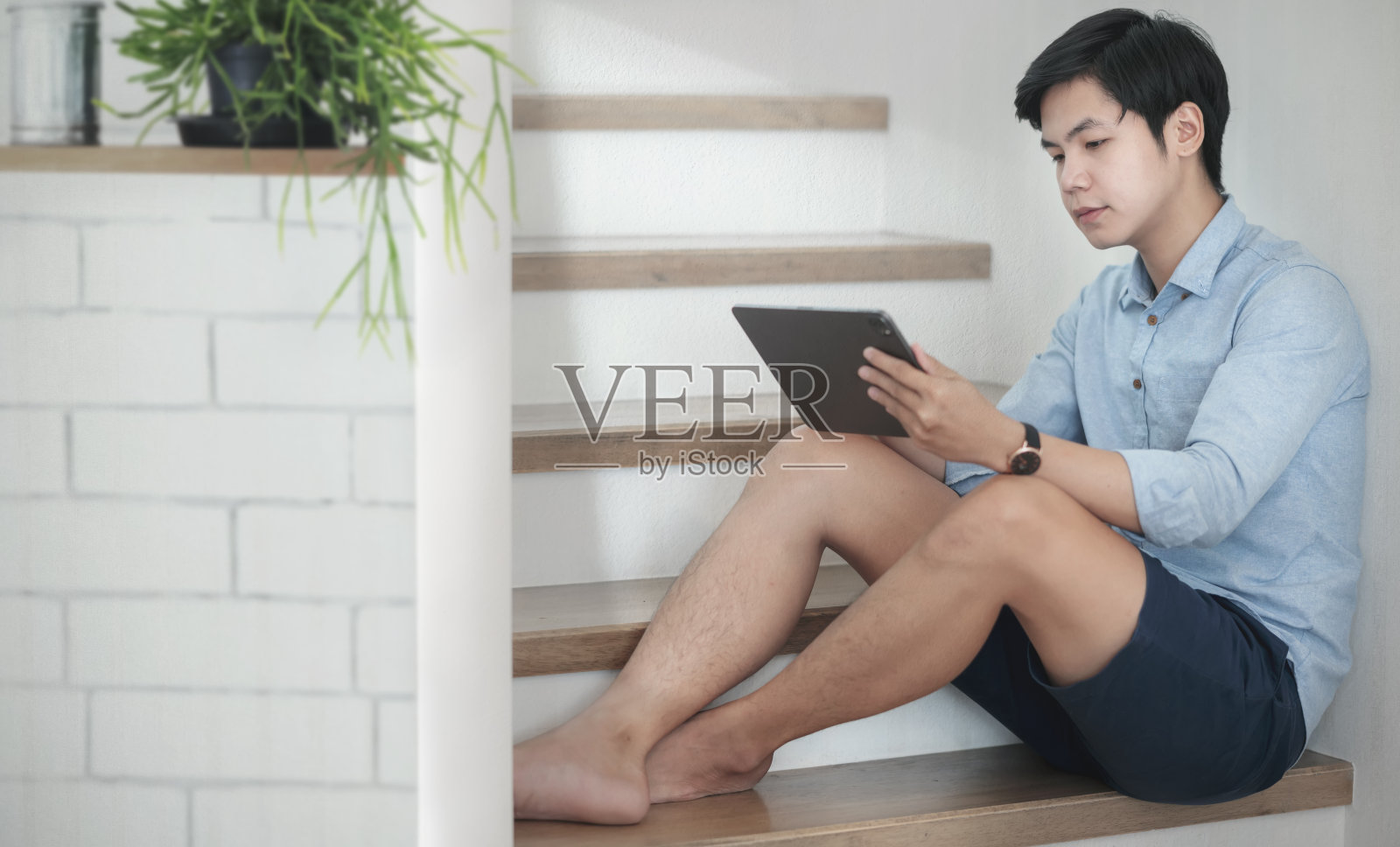 年轻的亚洲男子穿着休闲装，坐在家里的楼梯上使用平板电脑，复制空间。照片摄影图片