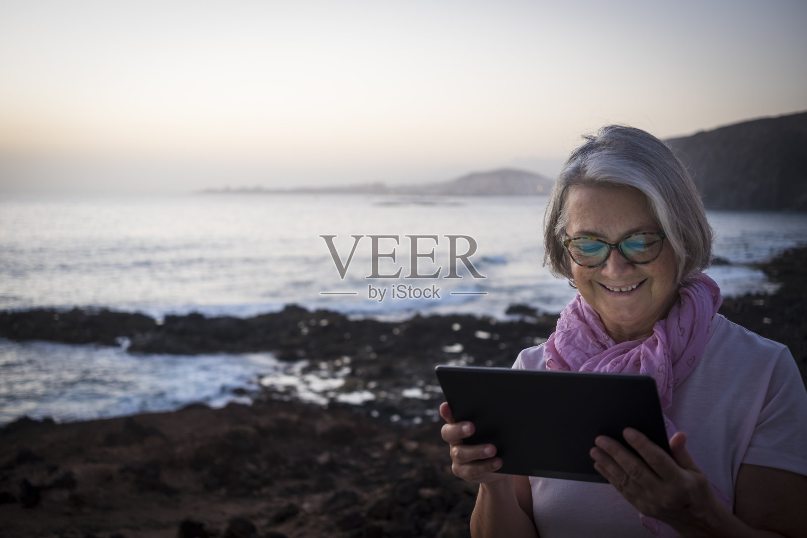 一位面带微笑的老年妇女坐在海滩附近，在黄昏的光线下看着平板电脑，与朋友进行视频通话。幸福的退休生活的概念。背景中的大海照片摄影图片
