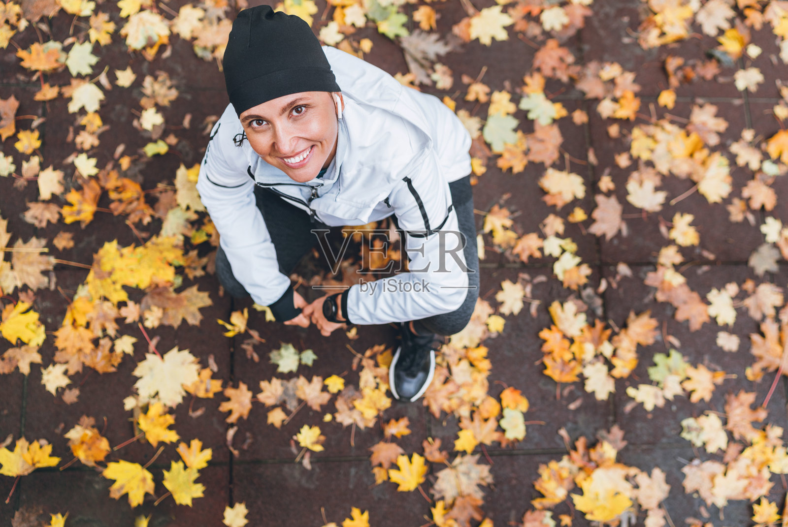 真诚微笑的白人女性跑步者休息后的早上慢跑，坐在城市公园的人行道上覆盖着潮湿的黄色秋天的叶子。活跃的运动人士俯视图概念形象。照片摄影图片