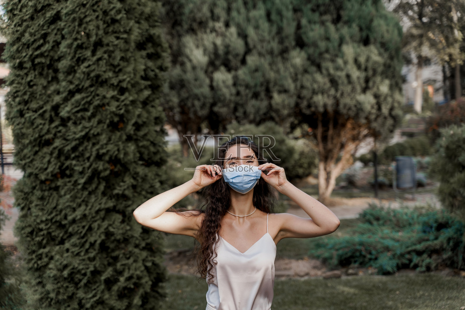 迷人女孩在covid-19隔离期接触医用口罩。在绿色的公园里，身着白色礼服的妇女。冠状病毒的概念照片摄影图片