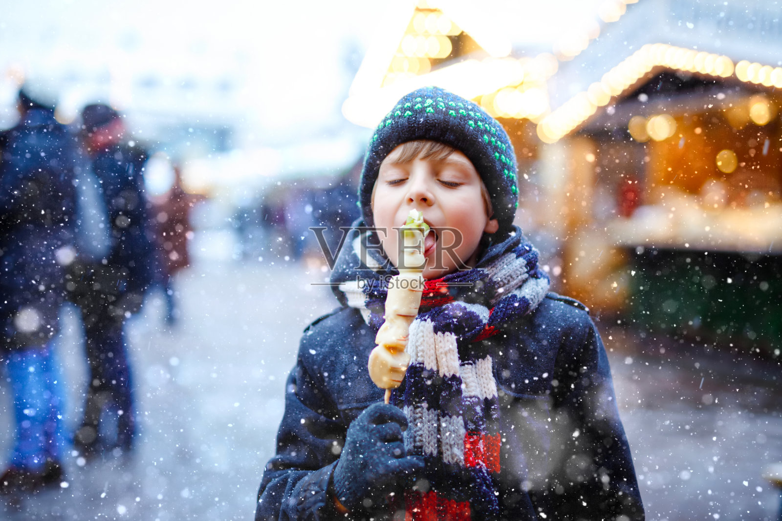 在德国传统的圣诞市场上，一个可爱的小男孩正在吃白巧克力串水果。快乐的孩子在传统家庭市场在德国雪天。照片摄影图片