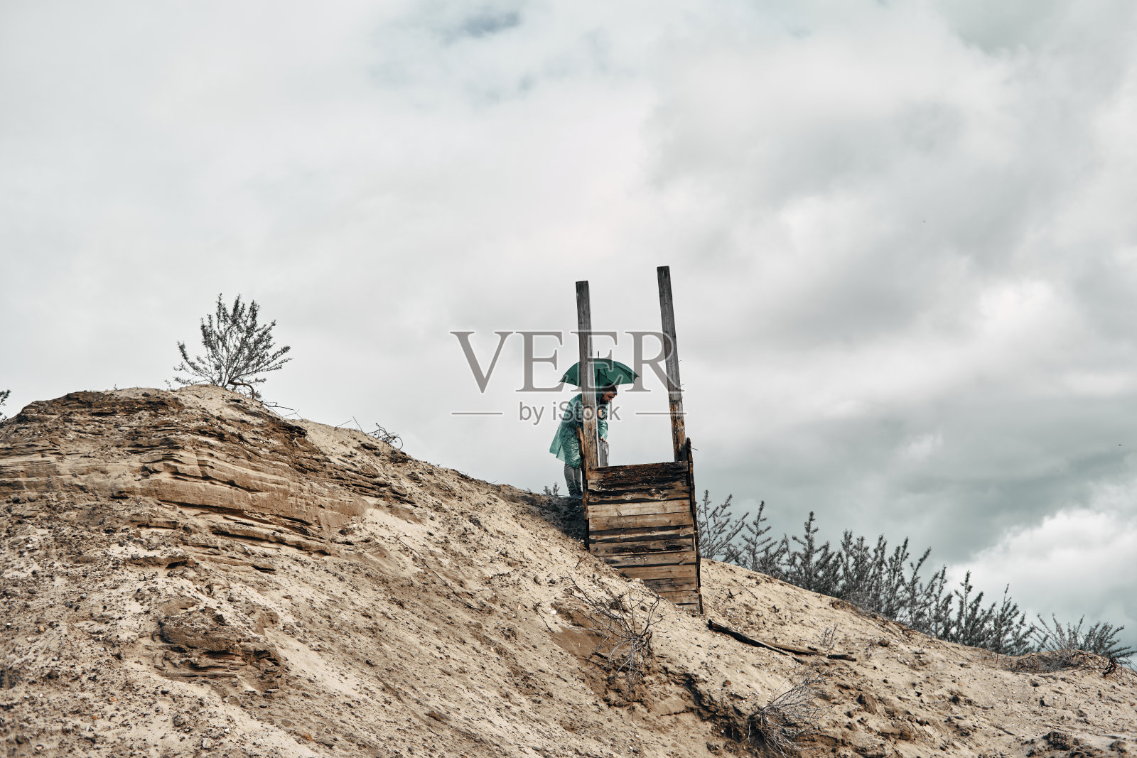 废弃的木井上的沙和一个人在井边穿着雨衣，天空多云。对废弃场所的研究。照片摄影图片
