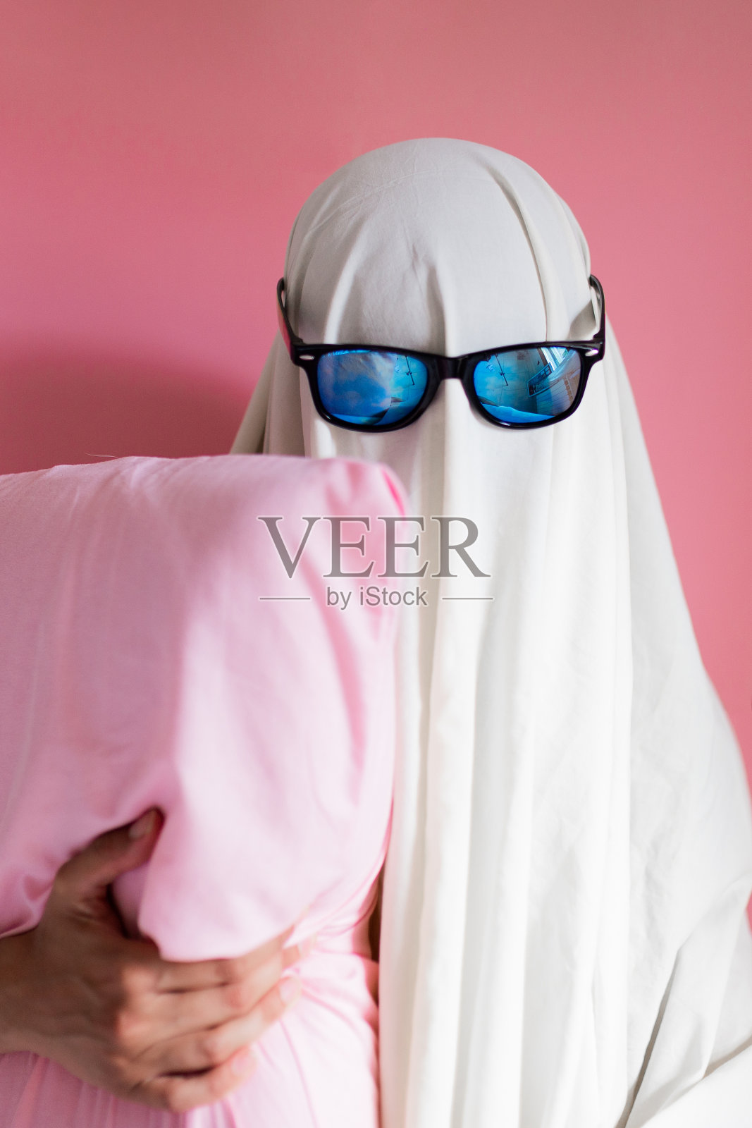 可爱的被单装扮鬼，戴着太阳镜，抱着粉红色的枕头。万圣节狂欢派对概念。照片摄影图片