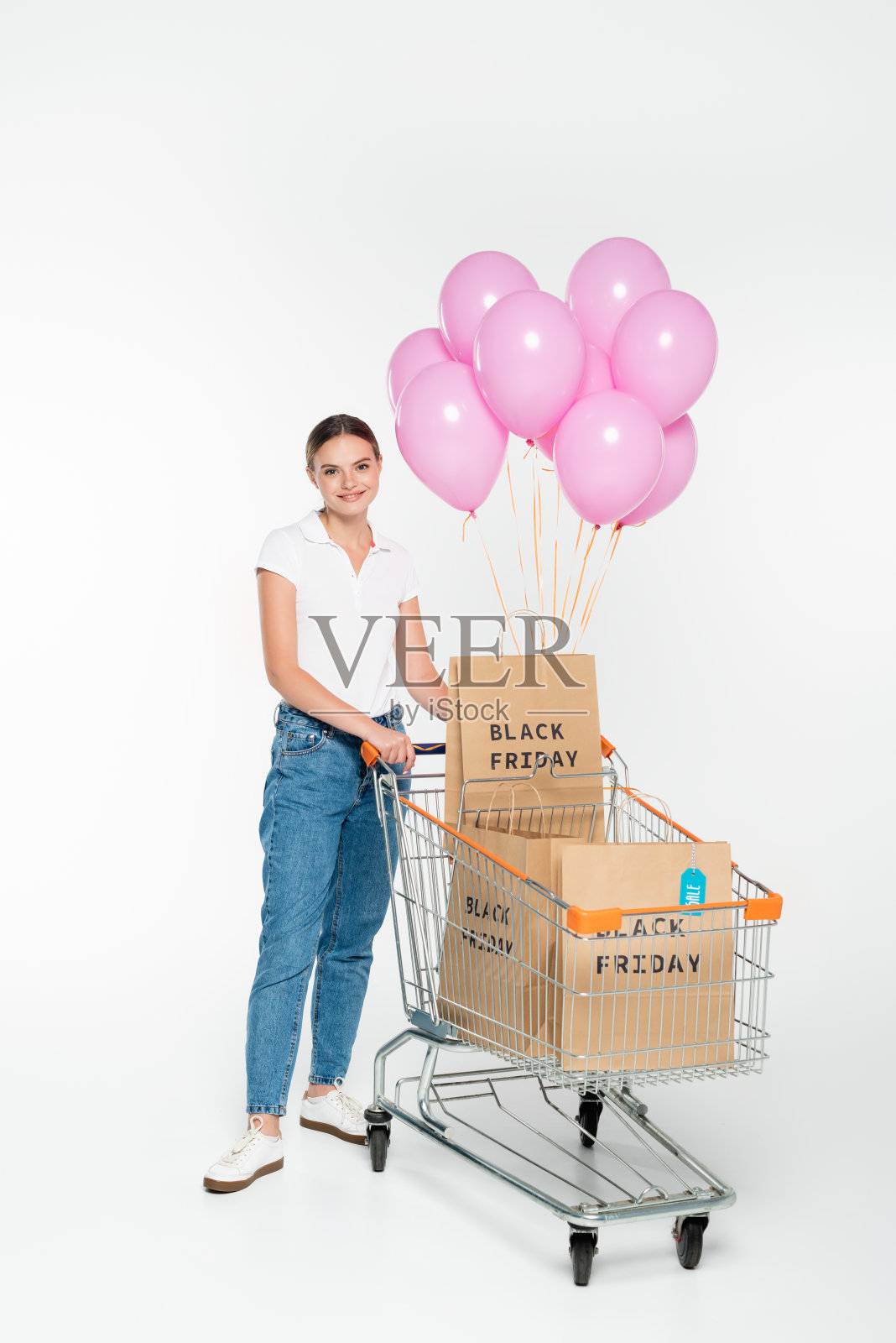 高兴的女人站在购物车旁边，拿着购物袋，黑色的星期五的字母和粉红色的气球在白色照片摄影图片