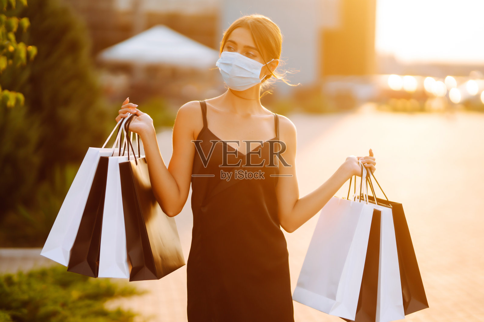美丽的女人戴着医用防护口罩，带着购物袋在日落时分走在购物中心附近。照片摄影图片