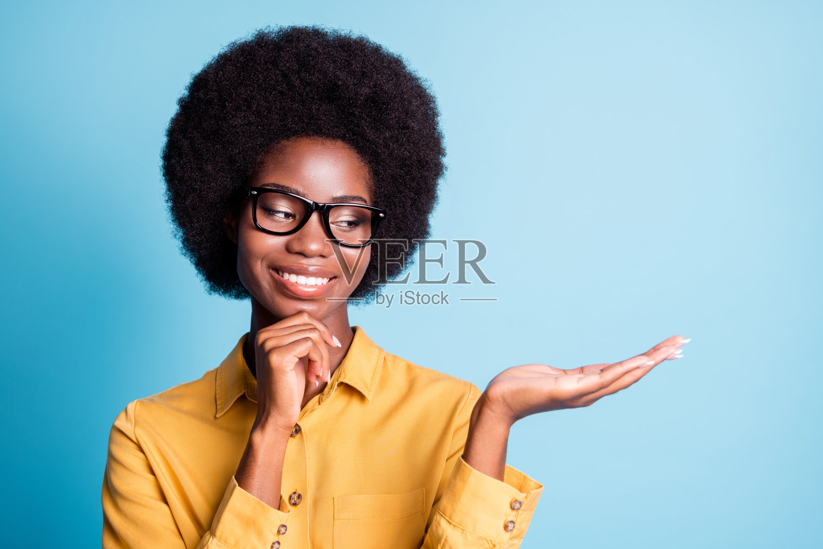 黑皮肤的照片，大体积的发型，女孩，闪亮的微笑，手，下巴，手掌，看空，认为有利的选择，穿着眼镜，黄色的衬衫，孤立的蓝色背景照片摄影图片