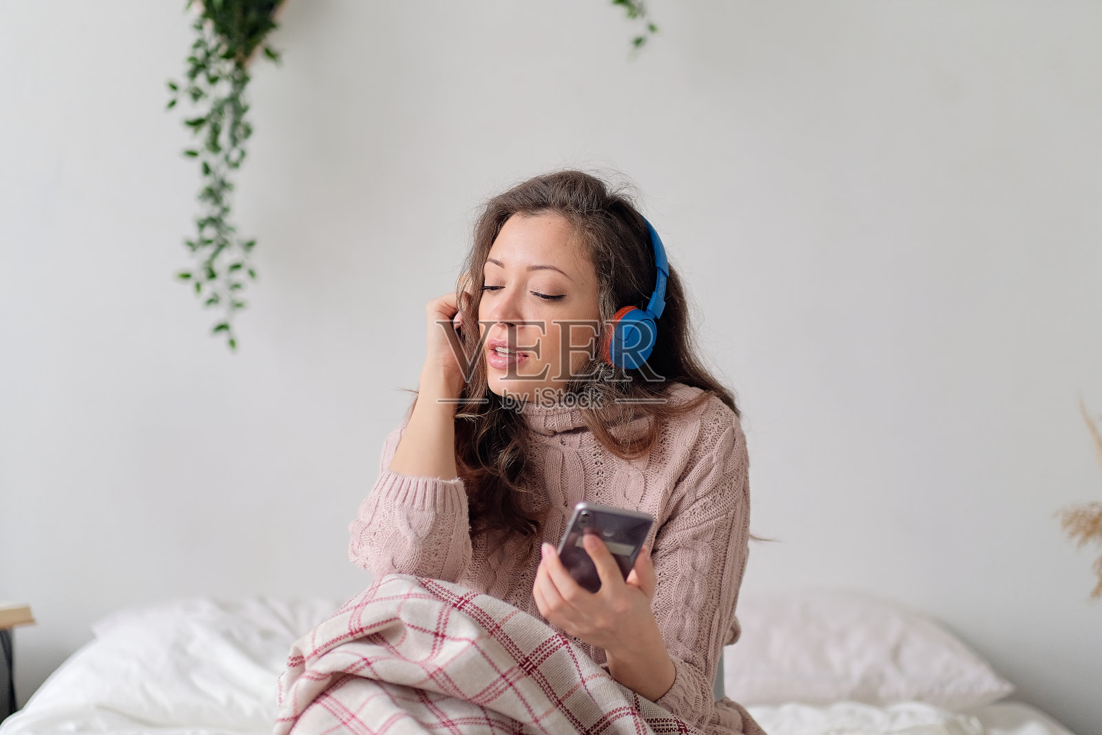 年轻快活的女人躺在床上，用手机拖着脚唱着歌。放松的女孩在家里用耳机听音乐。快乐的女人在不经意间享受着卧室里的音乐，同时发着手机短信照片摄影图片