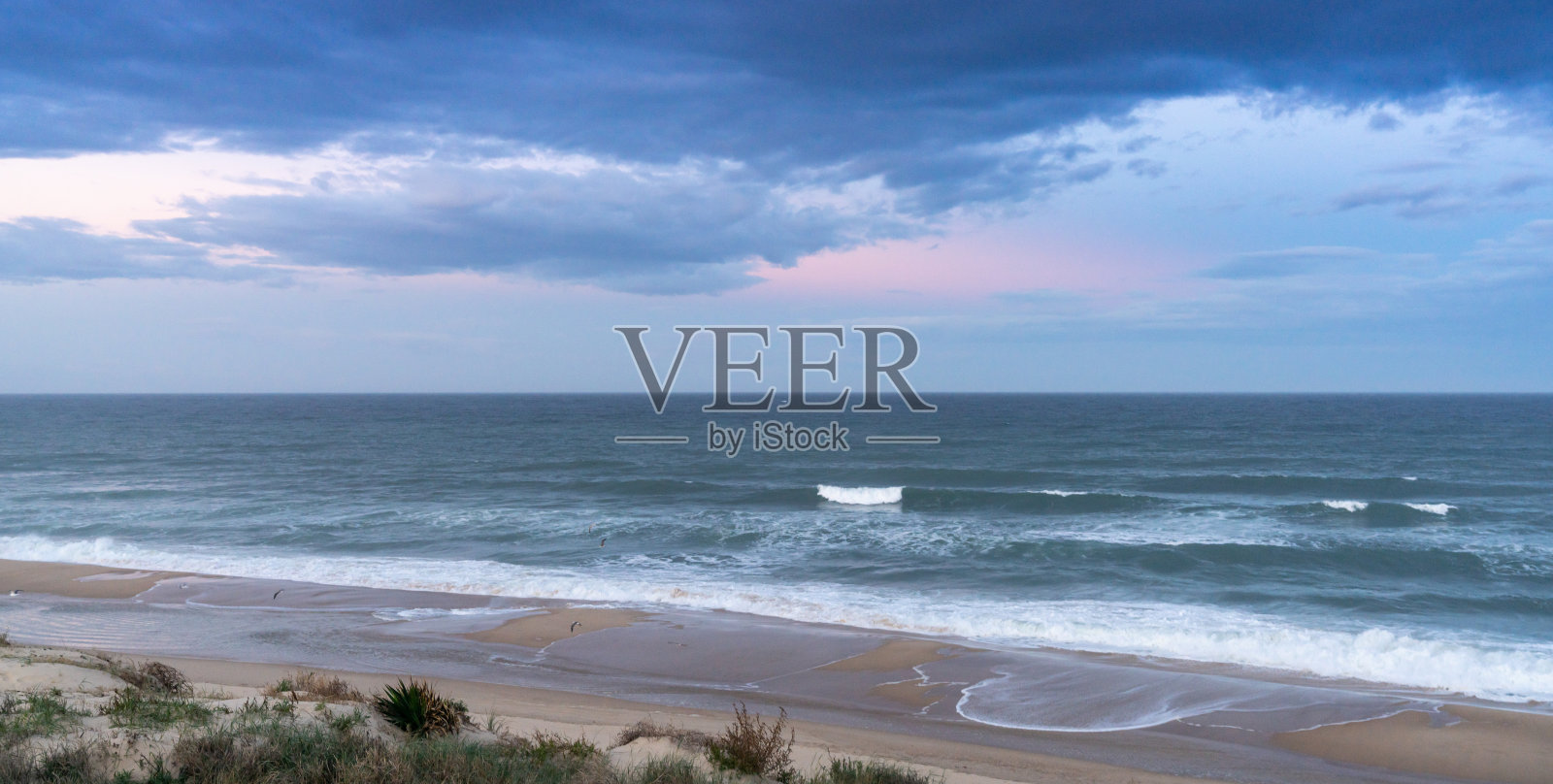在日出时暴风雨的天空下，沙丘和海浪拍打在无尽的海滩上照片摄影图片
