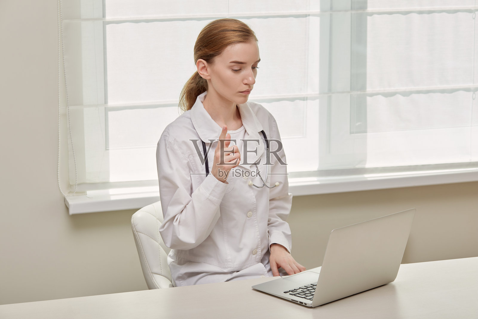 一位年轻的女医生，穿着医疗袍，看着笔记本电脑的屏幕，说着话。照片摄影图片