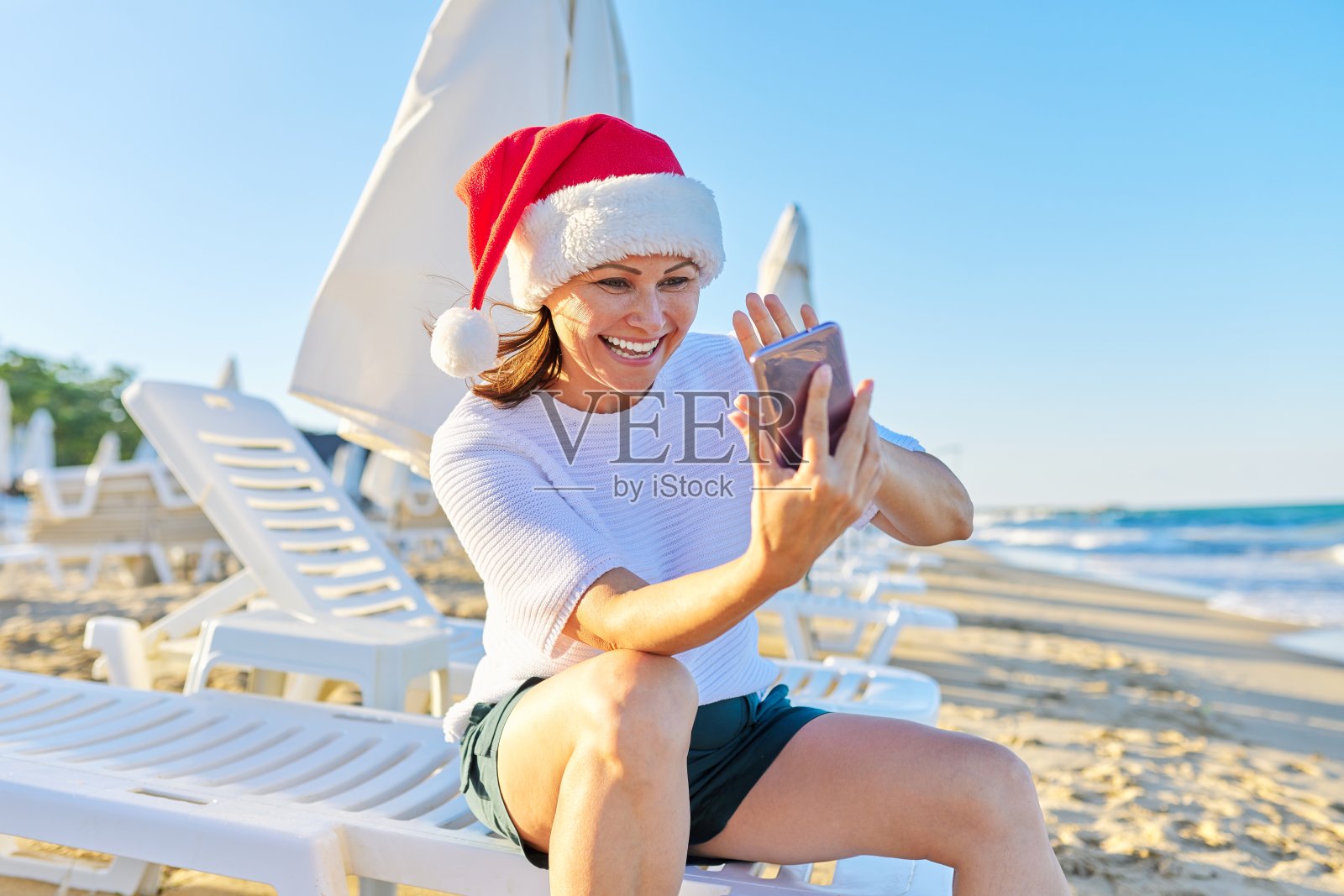 新年和圣诞假期，快乐的女人戴着圣诞帽在海滩上打视频电话照片摄影图片