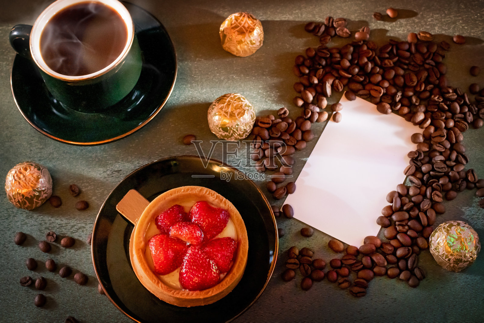 早餐有草莓蛋糕、香咖啡和巧克力。桌上的绿色台布上从工艺包里弄醒了咖啡豆。照片摄影图片