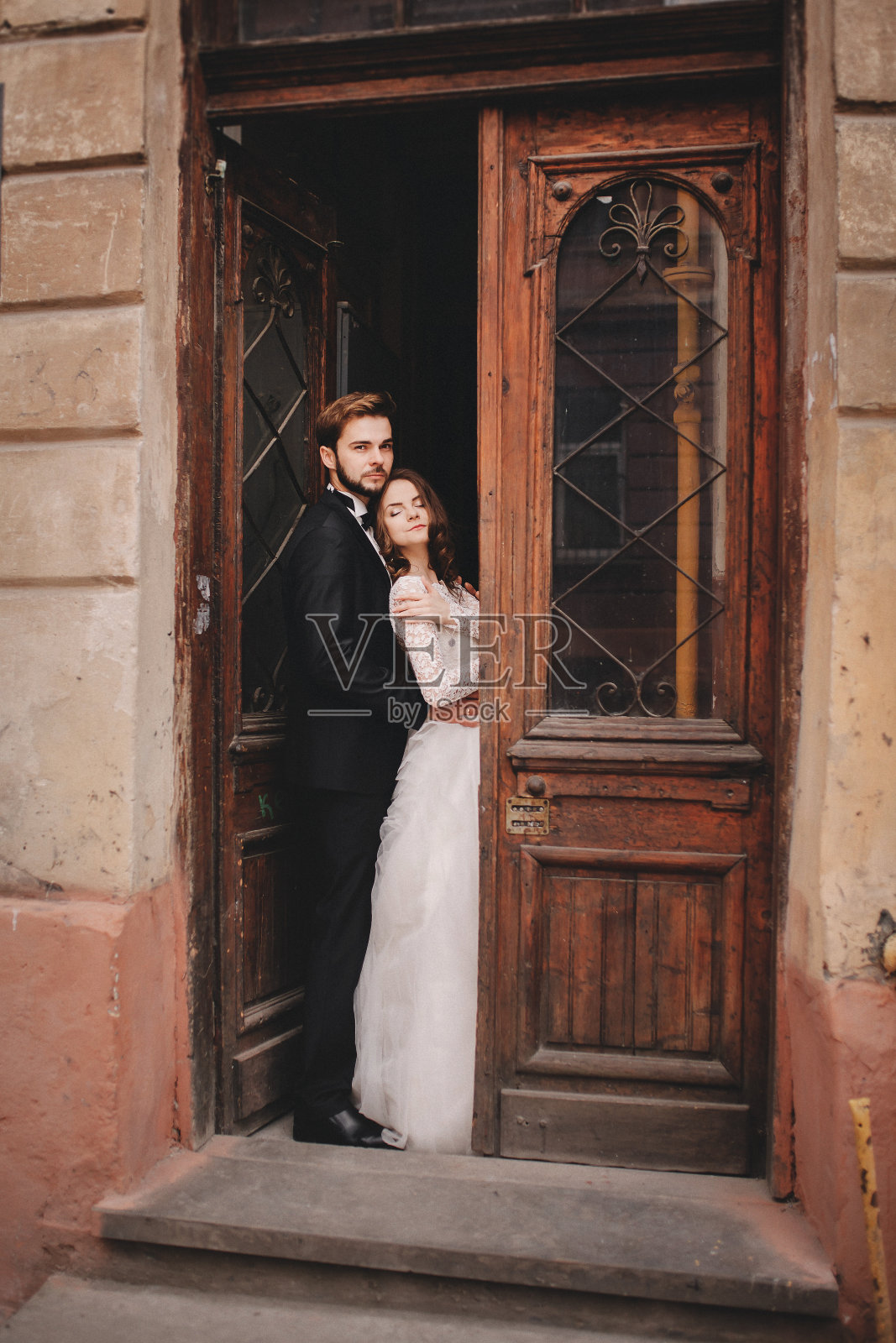 美丽的新婚夫妇拥抱在古老的门。一个时尚的新郎和一个年轻的新娘在一个欧洲小镇的老房子附近的婚礼肖像照片摄影图片