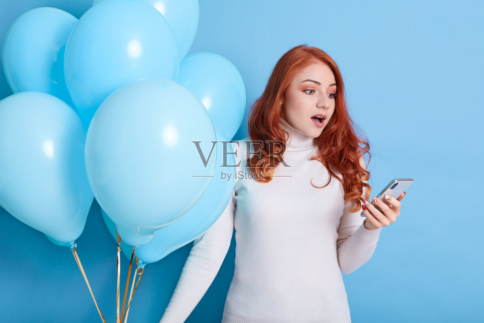 一名身穿白色毛衣和牛仔裤的年轻女子在蓝色背景下孤独地摆姿势庆祝生日，用手机举着气球，嘴巴张得大大的，表达着惊讶。照片摄影图片