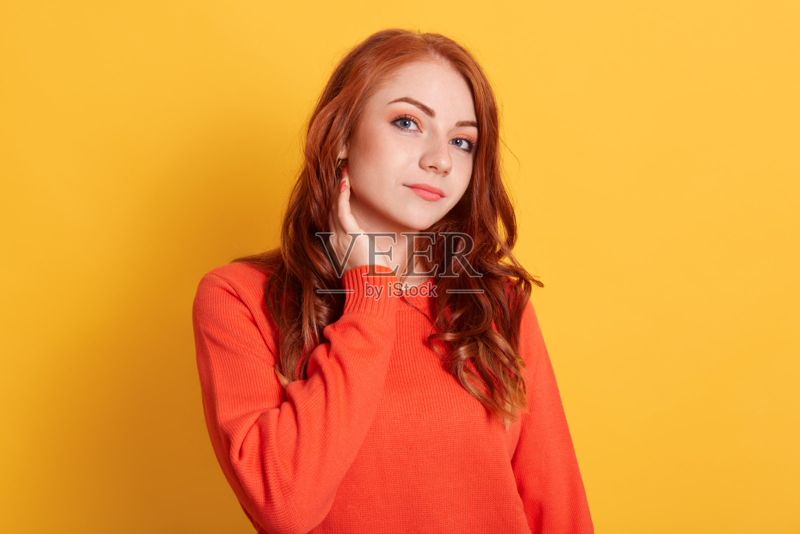 可爱的女人有美丽的眼睛，红色的长发，温柔的微笑，有愉快的表情和快乐，保持手在脖子上，穿着橙色的毛衣。照片摄影图片