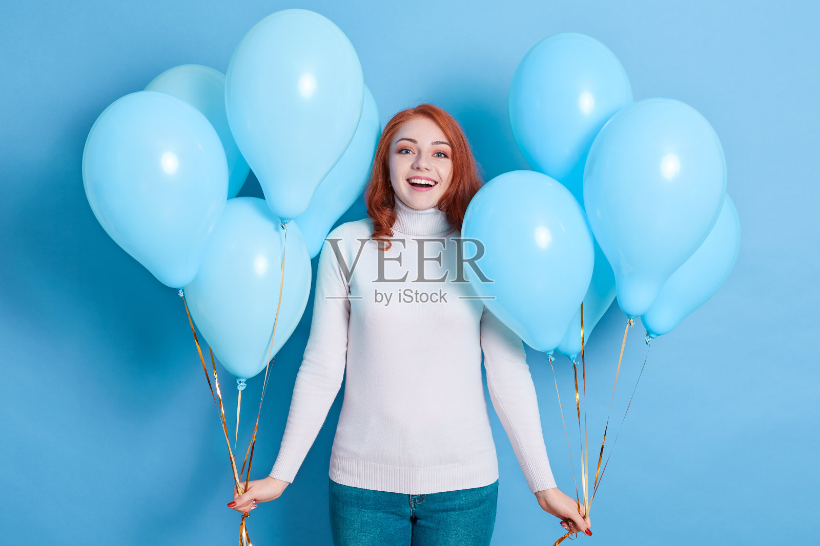 年轻女子女孩在毛衣和牛仔裤摆出孤立的蓝色背景，拿着两束气球，大张着嘴和兴奋的面部表情，看起来很高兴。照片摄影图片