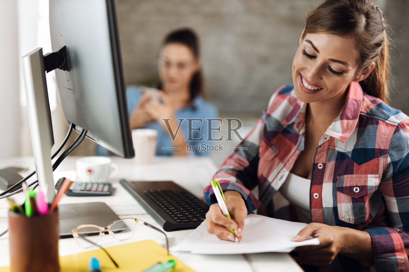 快乐的女商人一边在办公室用台式电脑工作一边做笔记照片摄影图片