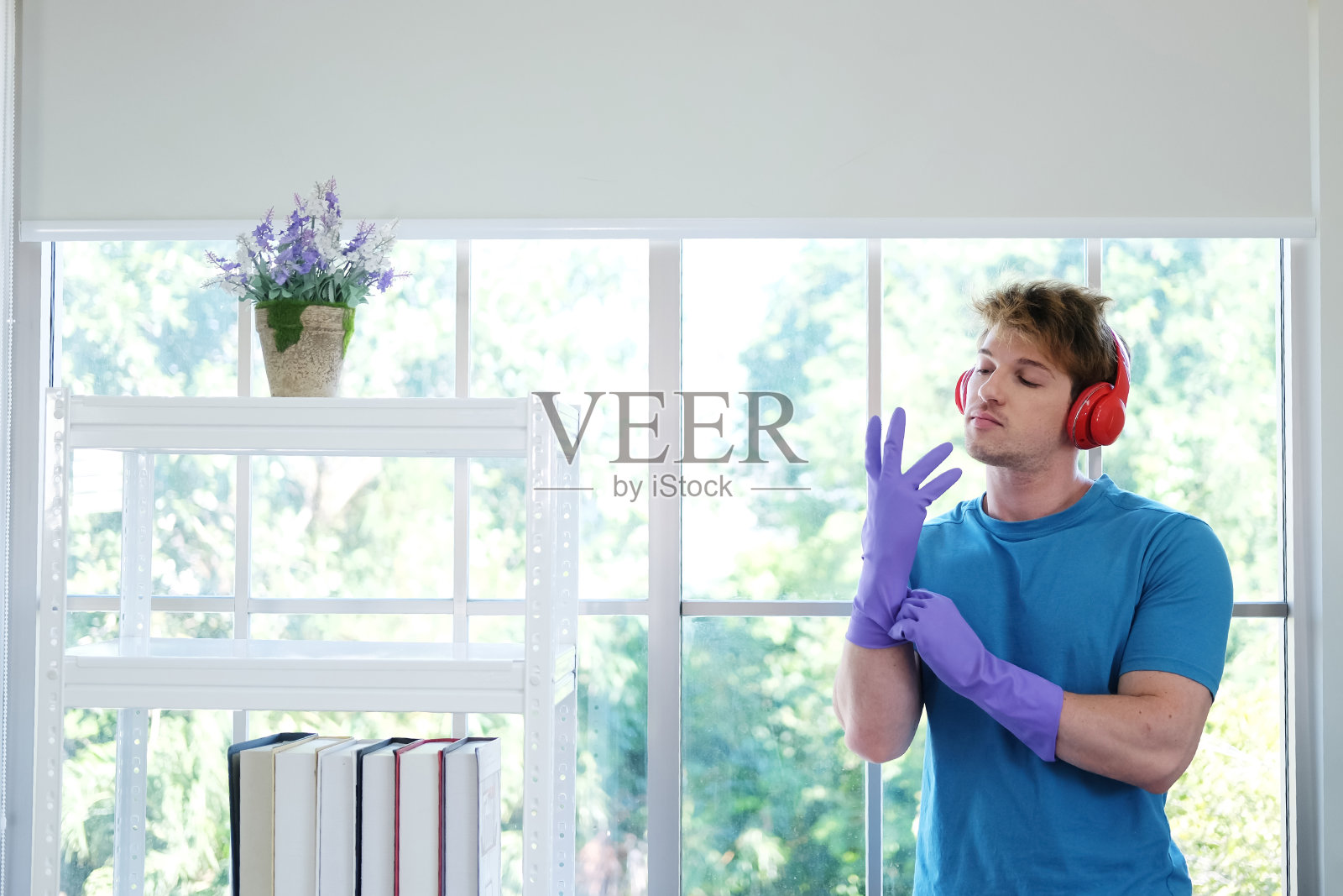 年轻的白人男子戴着紫色橡胶手套做家务，戴着红色耳机听音乐。家庭人的生活方式在假日概念。照片摄影图片