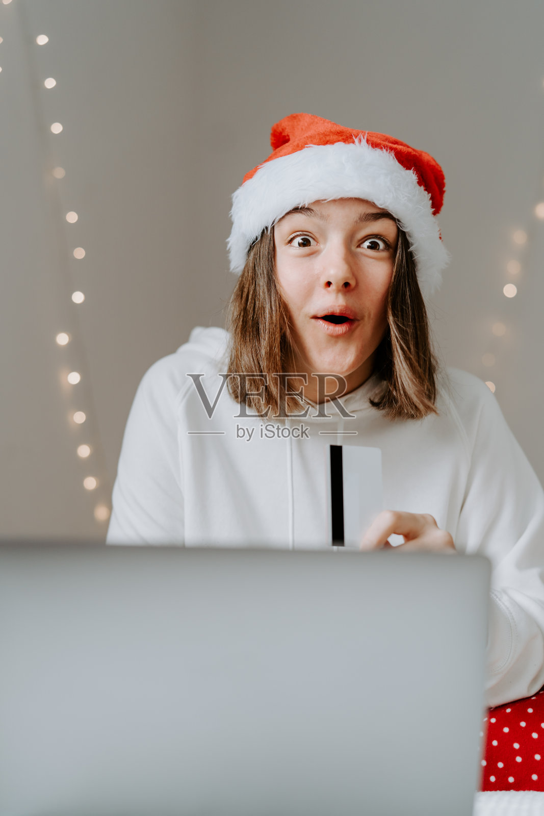 年轻快乐的十几岁的女孩兴奋的脸表情在红色的圣诞帽购物网上购买圣诞礼物用信用卡支付笔记本电脑看到大减价。照片摄影图片