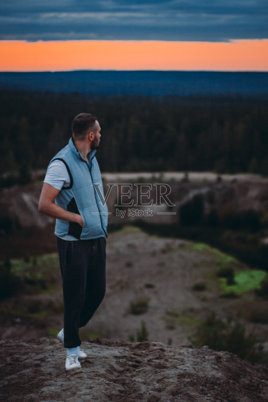 迷人的年轻人站在山上，看风景的森林和日落多云的天空照片摄影图片