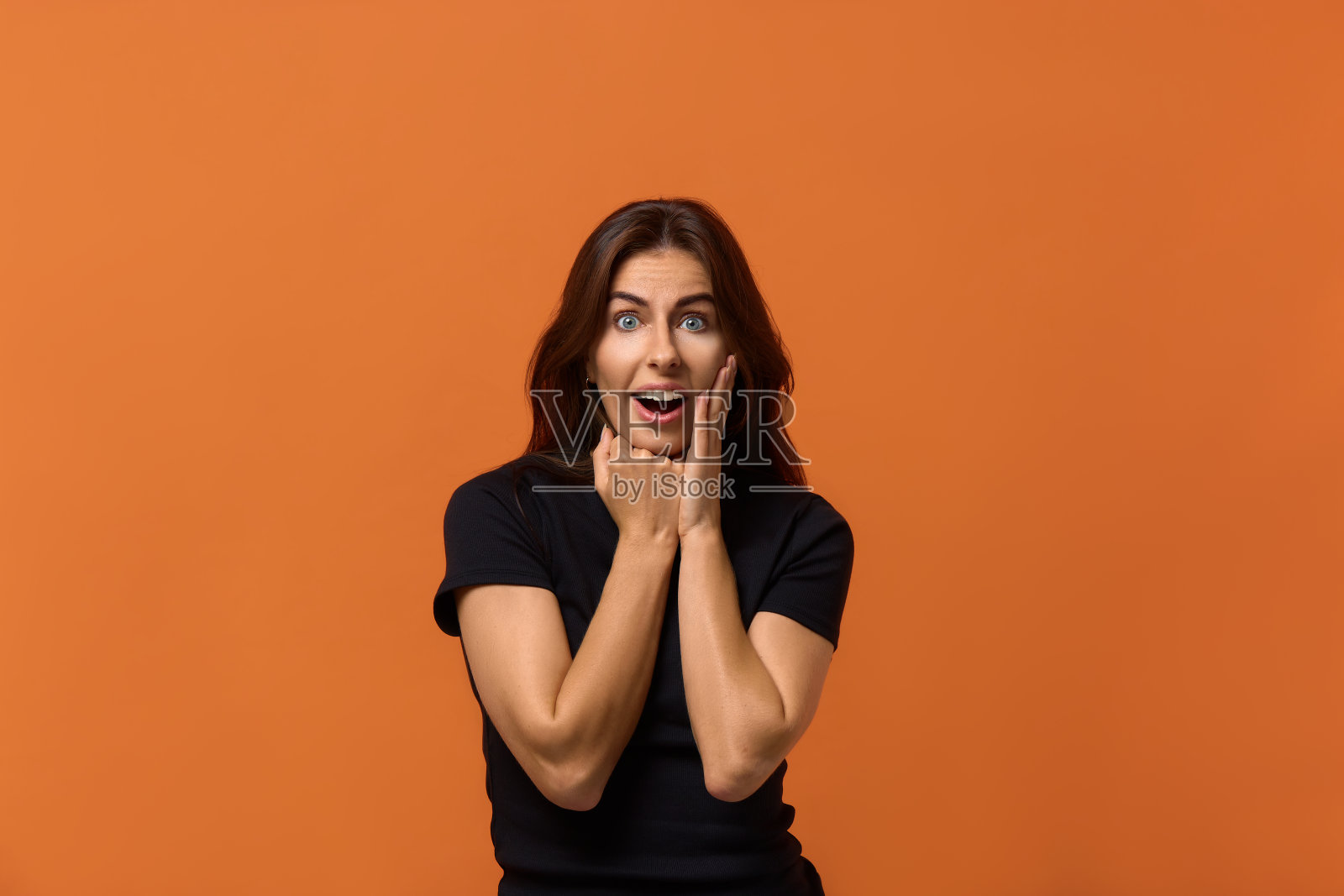 我真不敢相信。惊讶的白人妇女在黑色t恤拉手近脸在惊讶的手势，保持嘴和眼睛大开，看起来震惊。孤立在橙色背景上。照片摄影图片