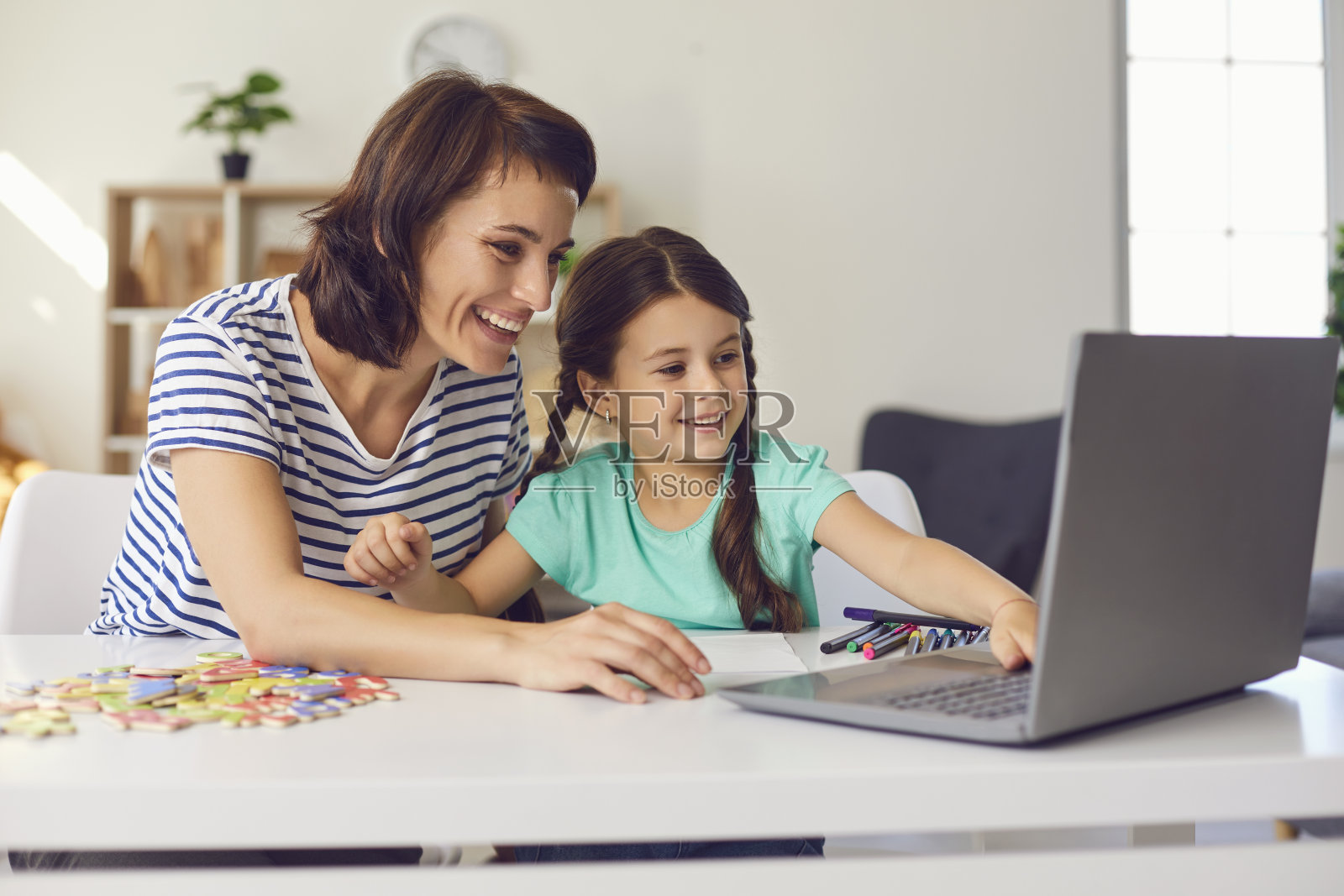 妈妈和她的小女儿笑着坐在桌子边看笔记本电脑上的视频。照片摄影图片