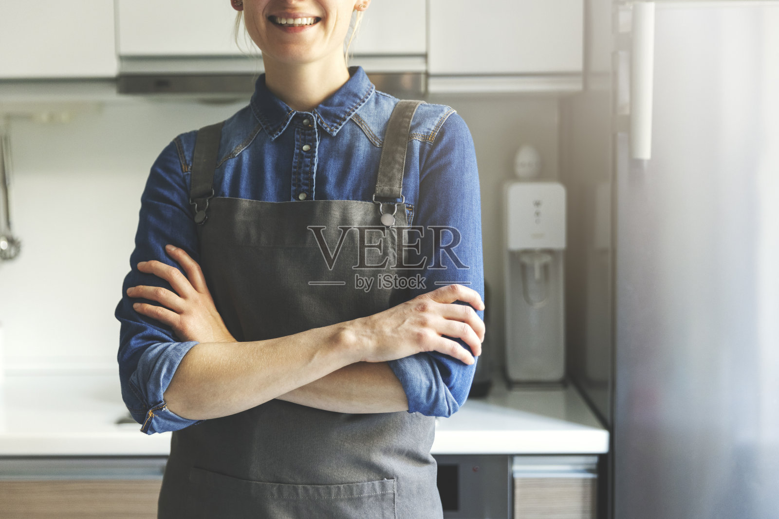 年轻的女人，穿着围裙，双手交叉站在厨房里照片摄影图片