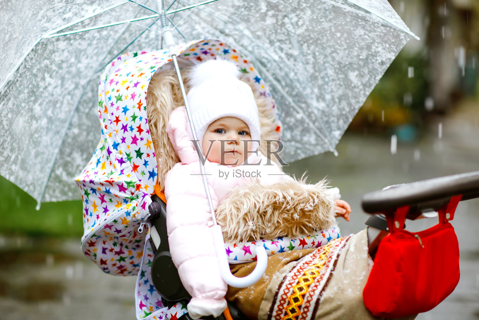 可爱的小女孩坐在婴儿车或婴儿车在寒冷的天，雨夹雪。快乐微笑的孩子穿着温暖的衣服，时尚时尚的婴儿外套。带着大伞的宝宝照片摄影图片
