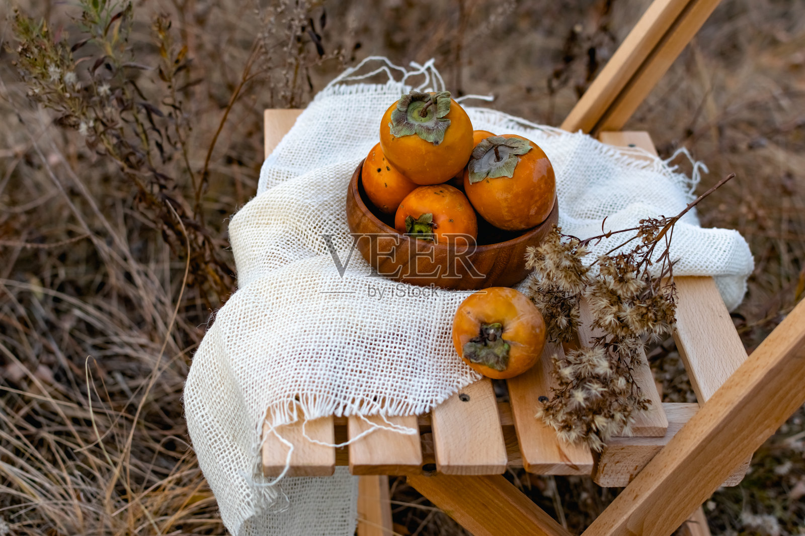 秋天组成的甜柿子品种，在一个木制盘子与干花序与一块粗麻布的木凳上。背景是树木的秋色照片摄影图片