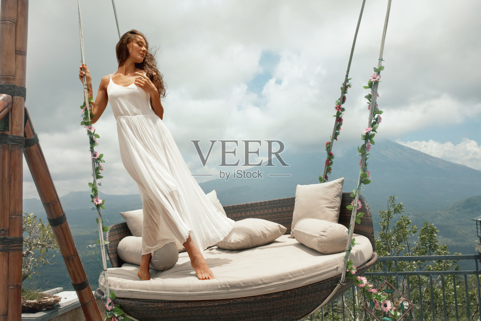 印度尼西亚巴厘岛，年轻女子身穿白色连衣裙，在阿贡火山前摆姿势。美丽的女孩站在挂椅上，山的背景是花。照片摄影图片