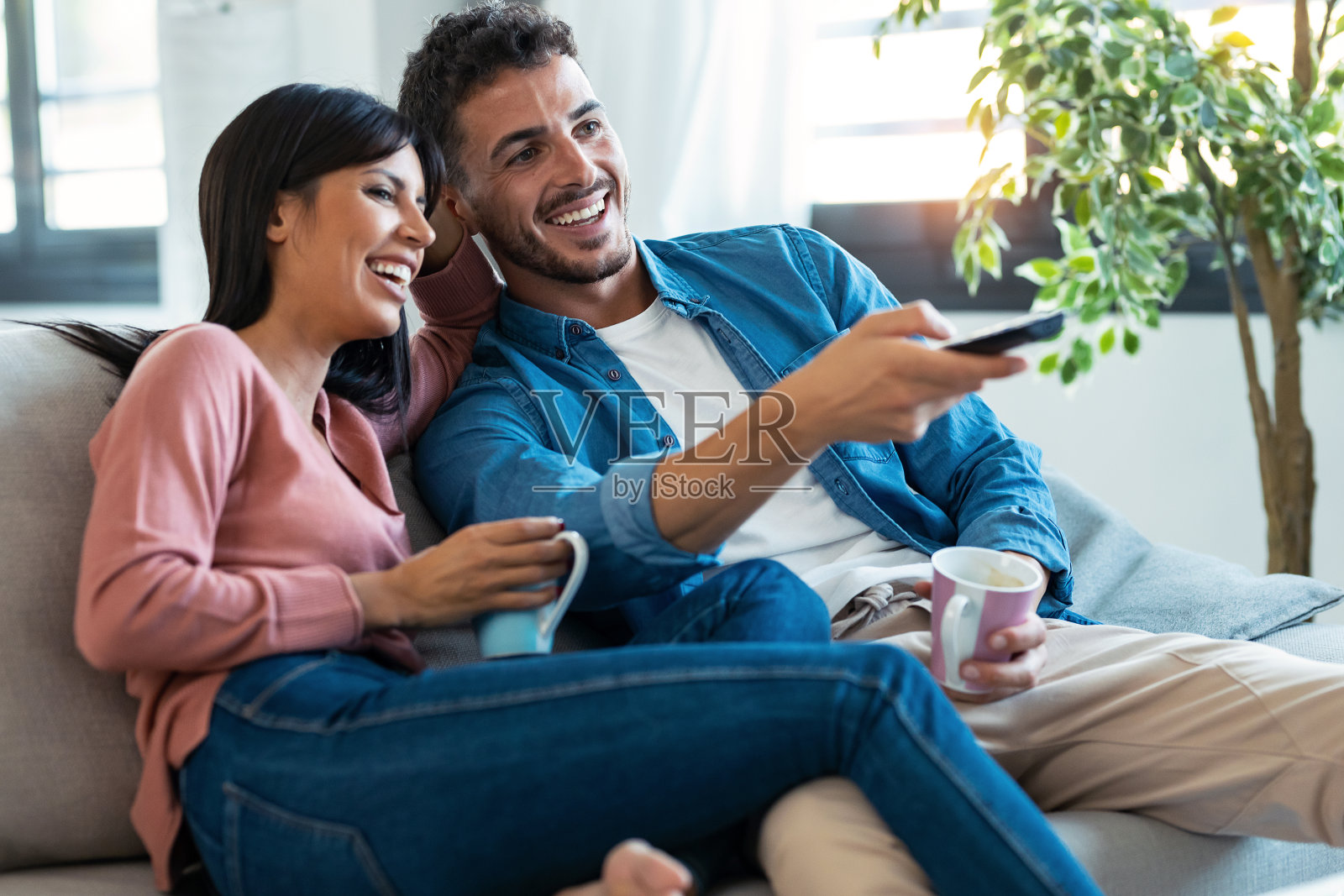 一对轻松的年轻夫妇一边坐在家里的沙发上看电视，一边用遥控器换频道。照片摄影图片