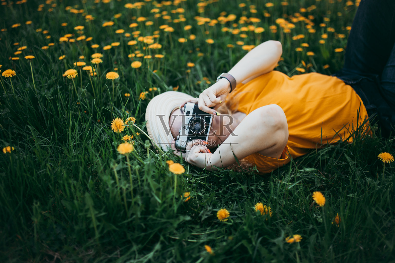 年轻美丽的女孩穿着橙色的t恤，手里拿着复古相机躺在蒲公英生长的草坪上，盛开的蒲公英照片摄影图片