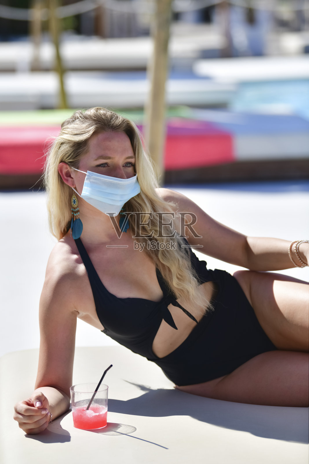 金发女人穿着泳衣和一个外科口罩躺在旁边的冷饮在一个模糊的背景。新的假期和安全理念。照片摄影图片