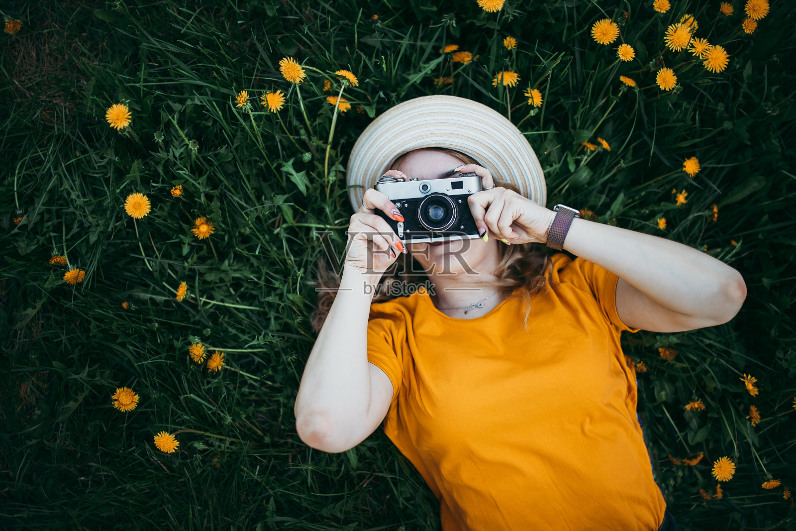 年轻美丽的女孩穿着橙色的t恤，手里拿着复古相机躺在蒲公英生长的草坪上，盛开的蒲公英照片摄影图片