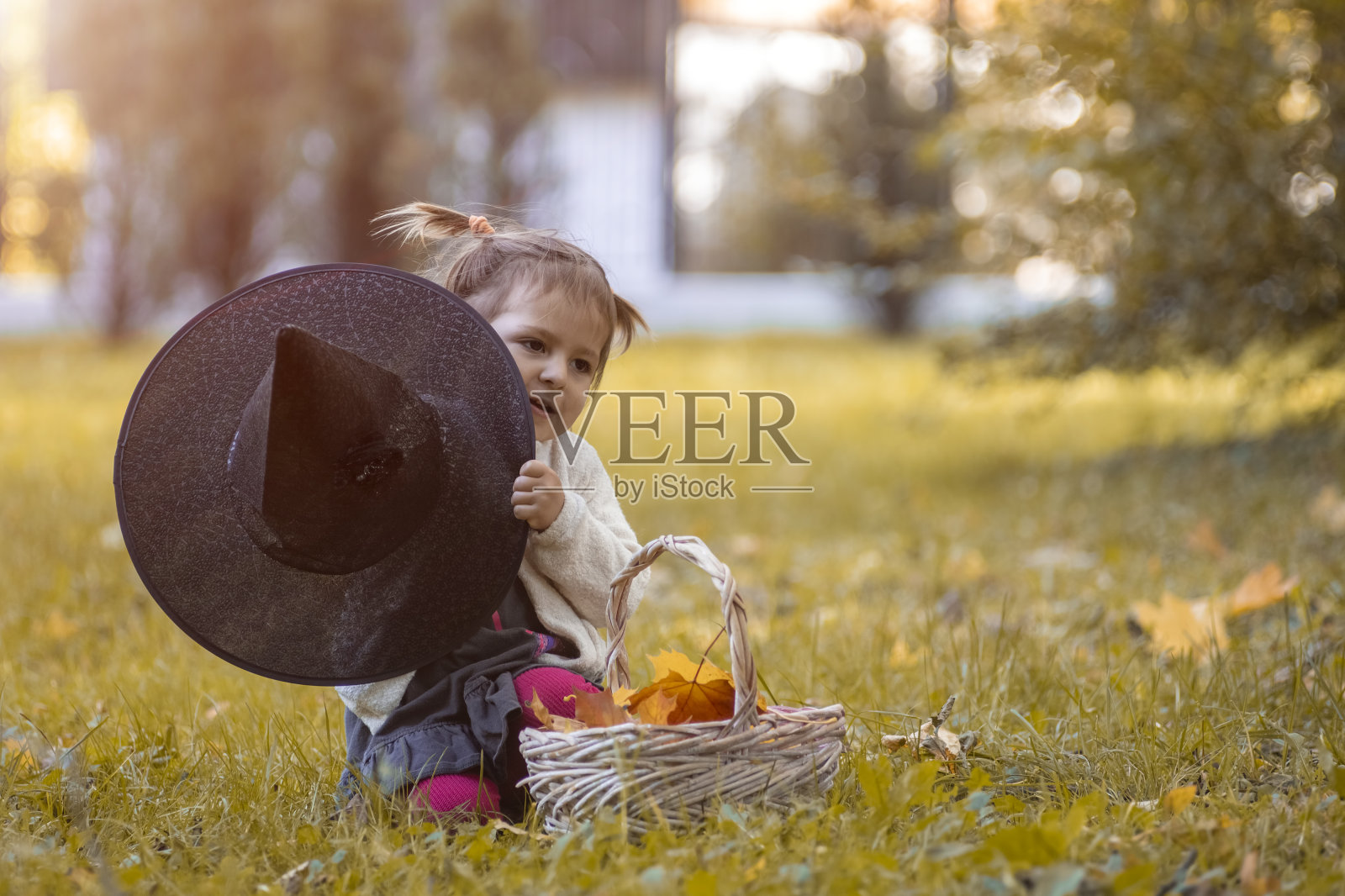 万圣节庆祝活动的概念。可爱的小孩在秋天的公园里躲在女巫的帽子后面玩耍。照片摄影图片