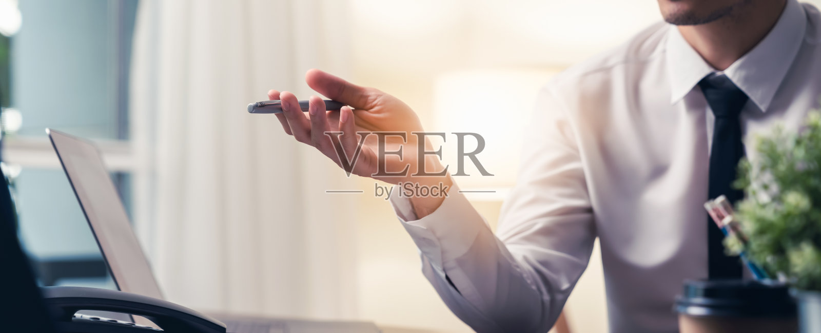 一个商人坐在桌子前，拿着一支笔指着前方。电脑和电话在现代办公室的桌子上。照片摄影图片