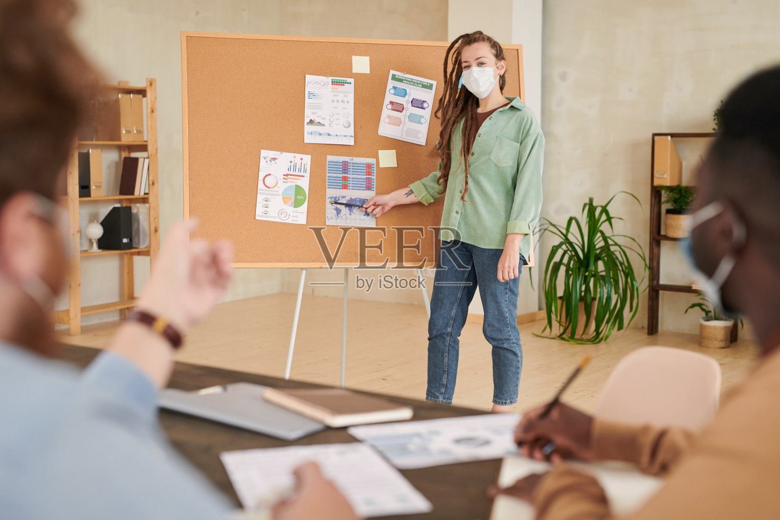 自信的女性营销人员戴着面具站在董事会上，指着纸上的图表，在会议上向同事们介绍策略照片摄影图片