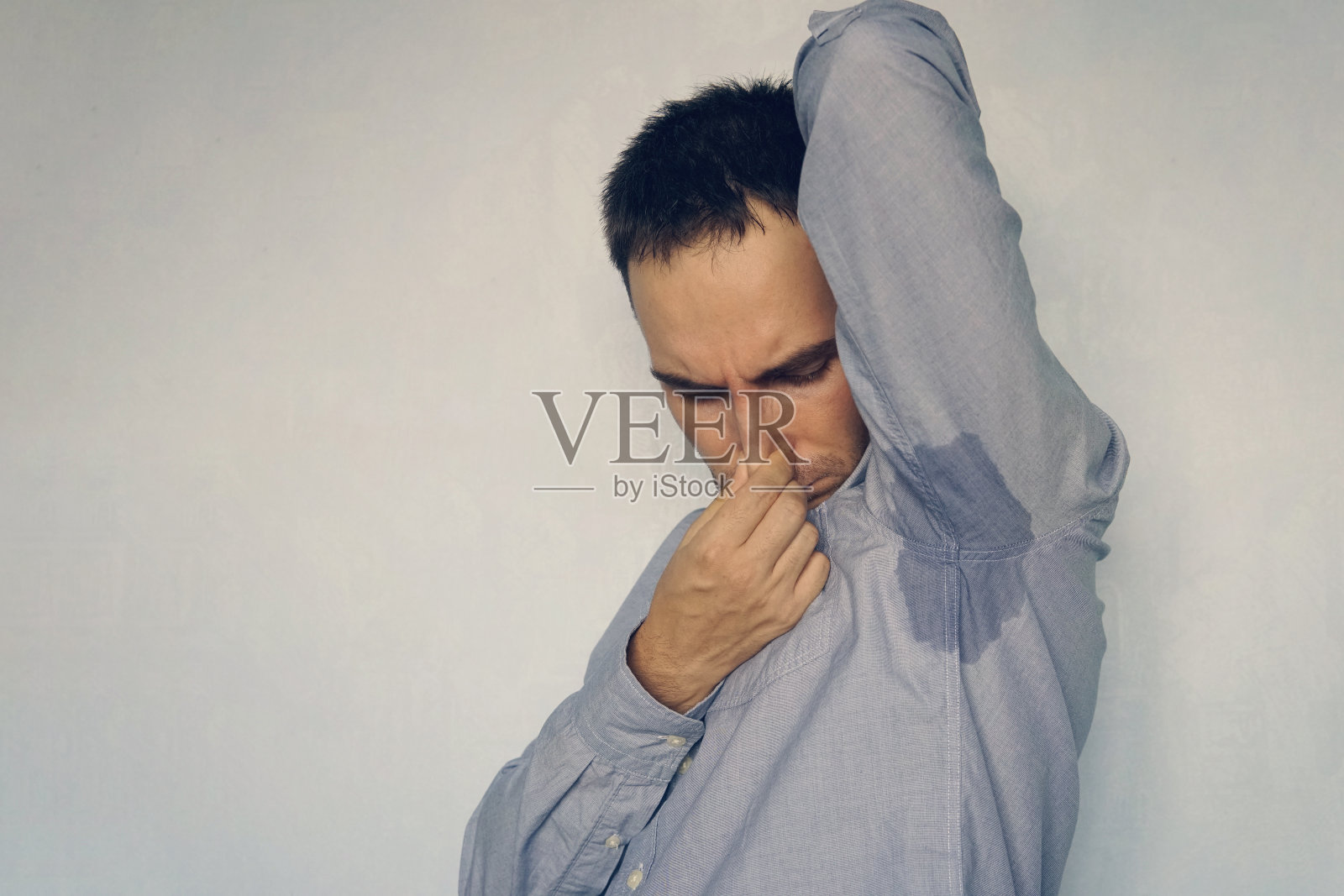 特写肖像年轻男子，嗅嗅腋窝，什么东西臭气熏天，恶臭的情况，蓝色的背景。消极的人类情绪，面部表情，感情照片摄影图片