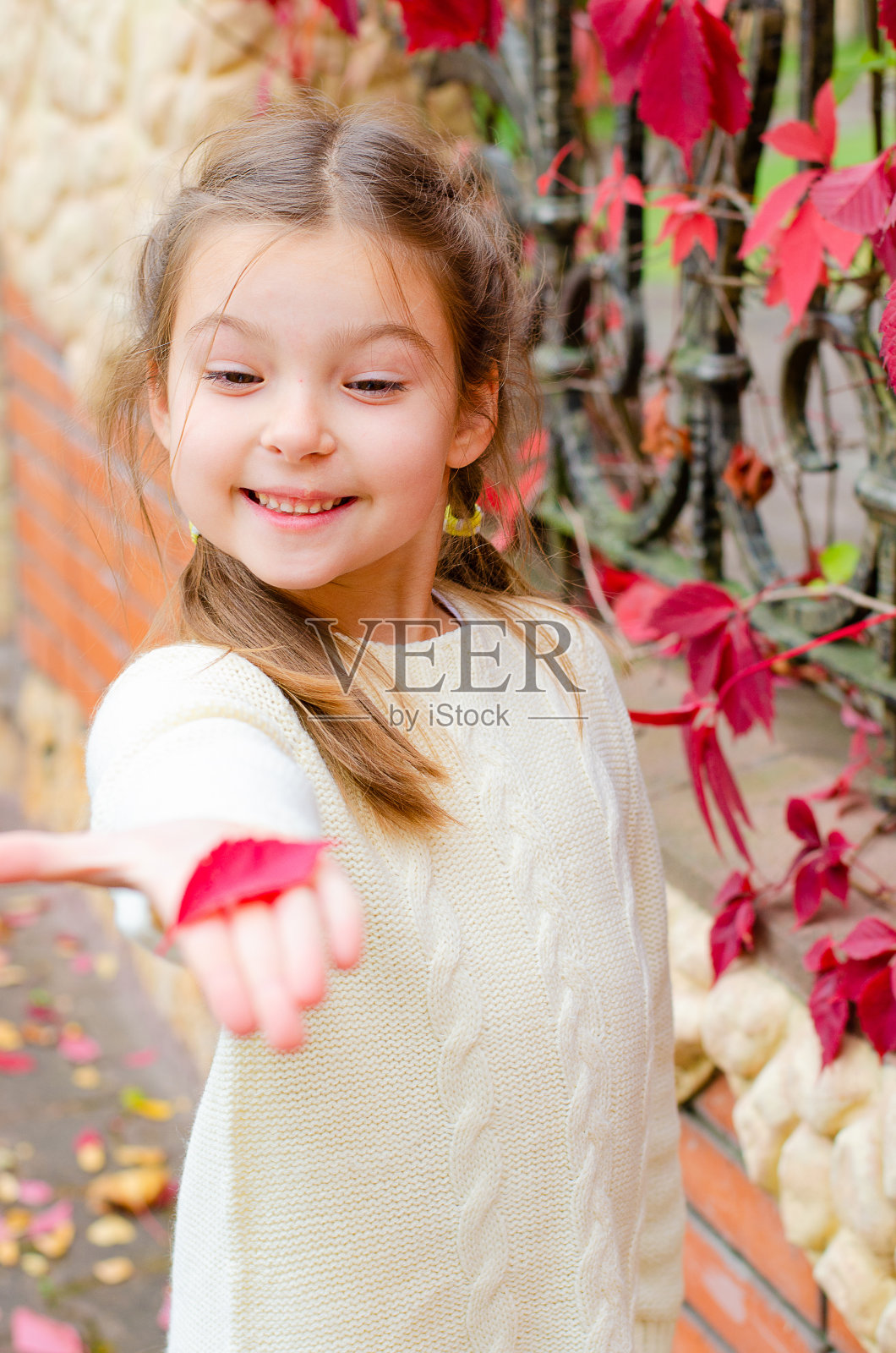 傍晚，在秋天的公园里，一个穿着白色针织连衣裙的小女孩，站在一堵有红绿葡萄叶子的墙旁。孩子微笑着，手里拿着树叶。照片摄影图片