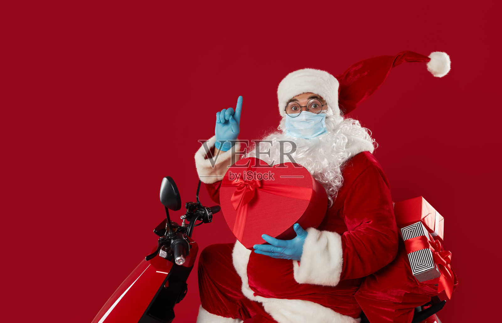 在冠状病毒隔离期间，圣诞老人戴着防护面具和手套，骑着复古摩托车递送一袋礼物。红色工作室背景。照片摄影图片