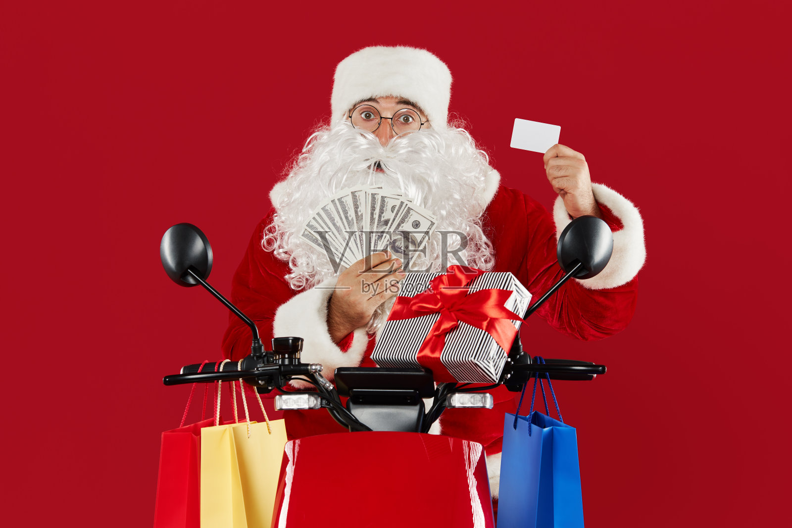 一个骑着复古摩托车的快乐疯狂的圣诞老人的肖像。送礼物在一个红色的工作室背景。使用信用卡通过手机进行在线订购。圣诞节和新年购物照片摄影图片