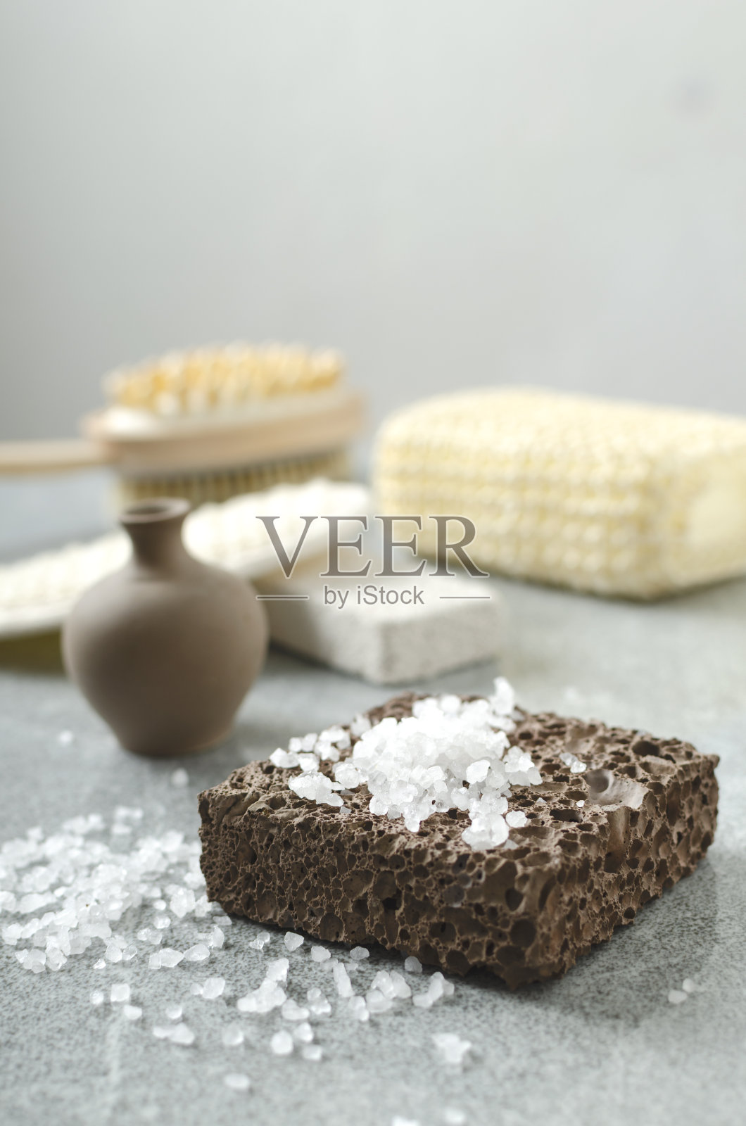 垂直的图像。棕色的石头和用于擦洗的海盐的特写，一瓶香油和不同的海绵放在灰色的桌子上，靠在明亮的墙壁上照片摄影图片