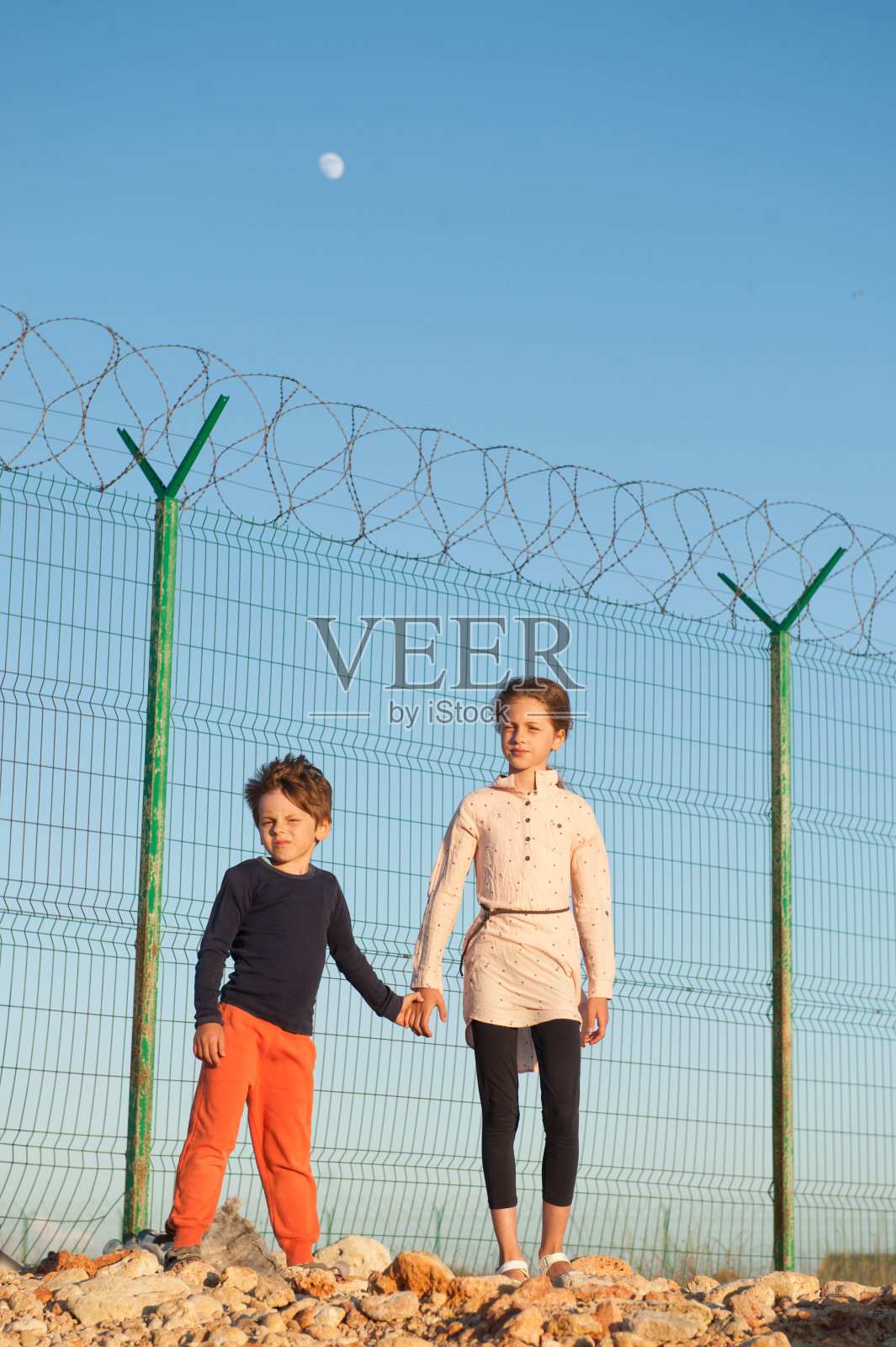 希望自由的小儿童难民家庭移民孤儿小男孩哥哥牵着姐姐女孩的手与关怀和爱站在石头与铁丝网和蓝天与月亮照片摄影图片