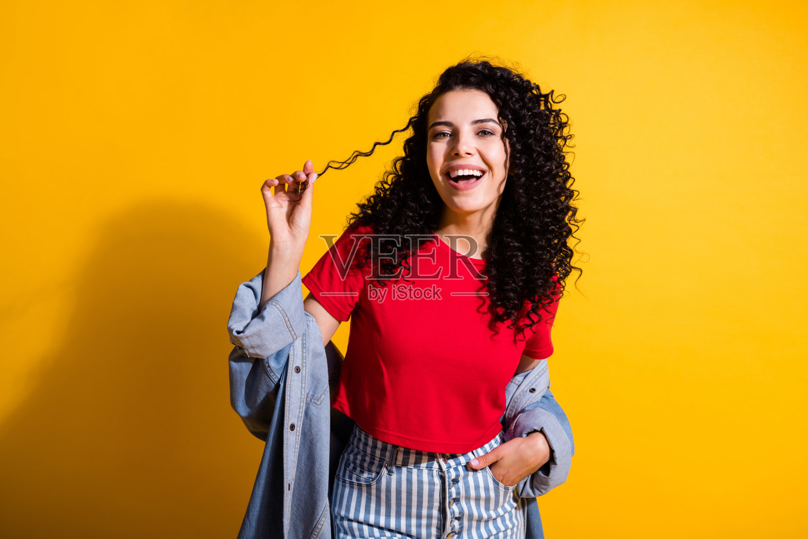 可爱的女孩玩卷发，张开嘴，手口袋，穿红色上衣条纹牛仔裤，夹克孤立的黄色背景照片摄影图片