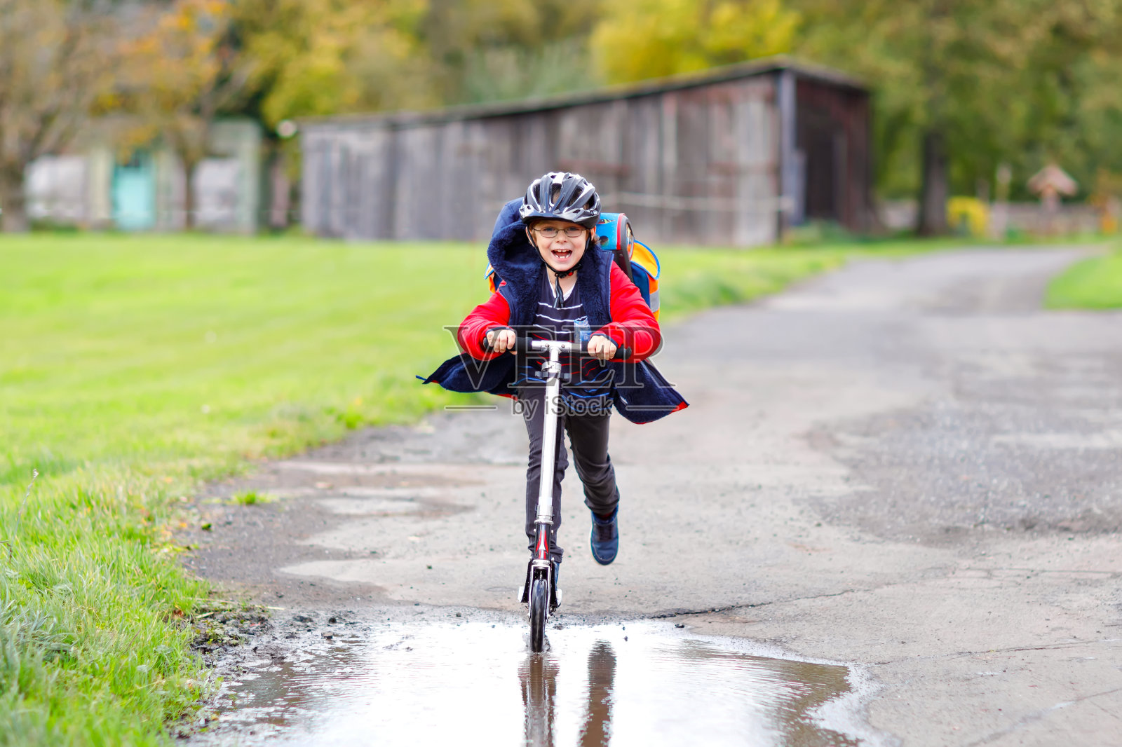 可爱的小男孩骑着滑板车在往返学校的路上。7岁的小男生开车穿过雨坑。有趣的快乐的孩子在五颜六色的时装和头盔。照片摄影图片